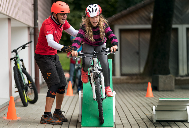 Kind fährt mit dem Fahrrad über eine Rampe, ein Erwachsener leistet Hilfestellung. 