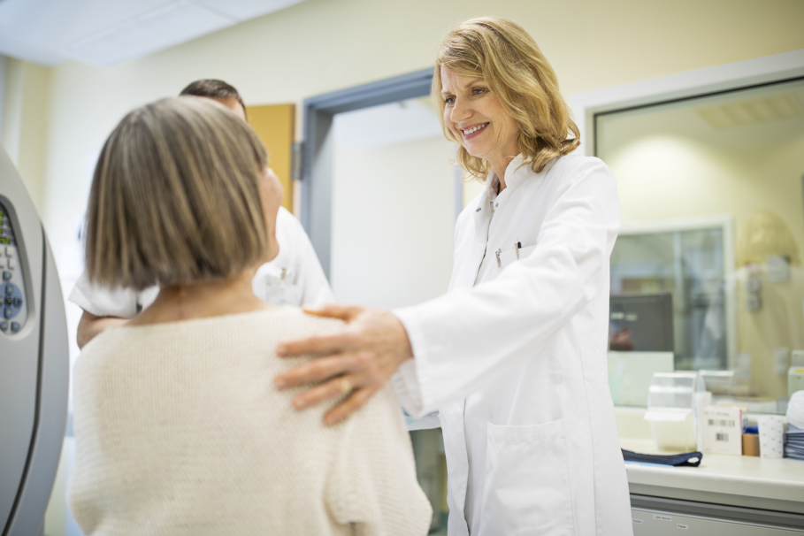 Patienten-Ärzte-Situation: Die Ärztin steht und lächelt die sitzende Patientin an, ihre linke Hand drückt die linke Schulter der Patientin. 