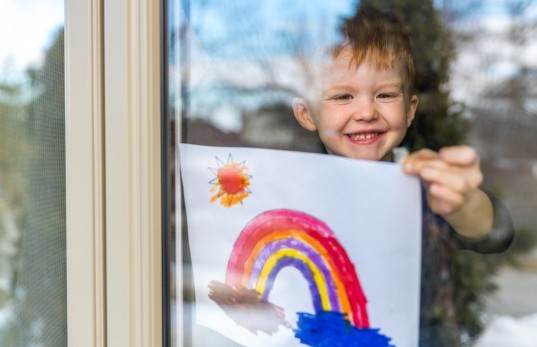 Kind steht am Fenster, blickt nach draussen und hält lächelnd einen gemalten Regenbogen gegen die Scheibe.